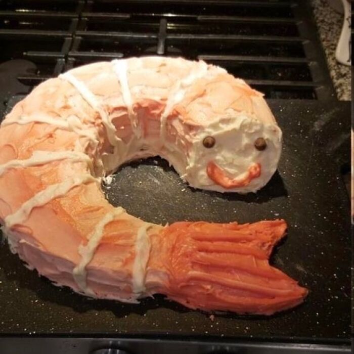 Shrimp-shaped cake with a smiling face, decorated with pink and white frosting, featured in relatable memes collection.