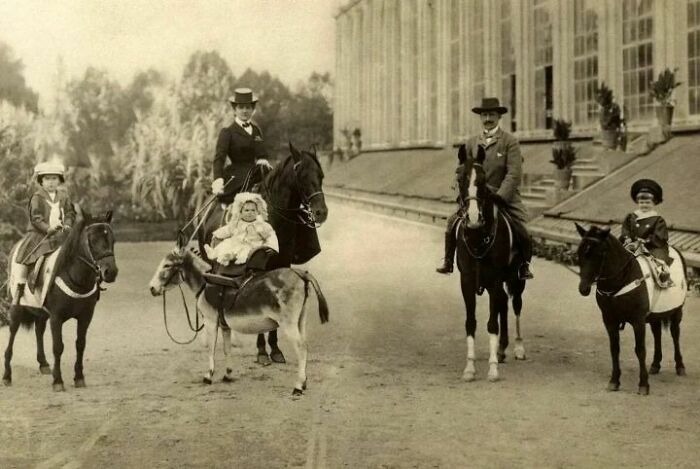 Vintage photo showing a family dressed in period clothing riding horses and a donkey outside a grand building.