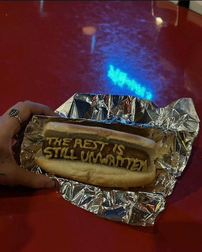 Hot dog with mustard text reading the rest is still unwritten, relatable memes on foil wrapper over red surface.