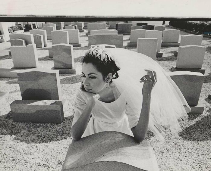 Bride in vintage wedding dress and veil sitting among tombstones in a cemetery, capturing a mesmerizing vintage photo.