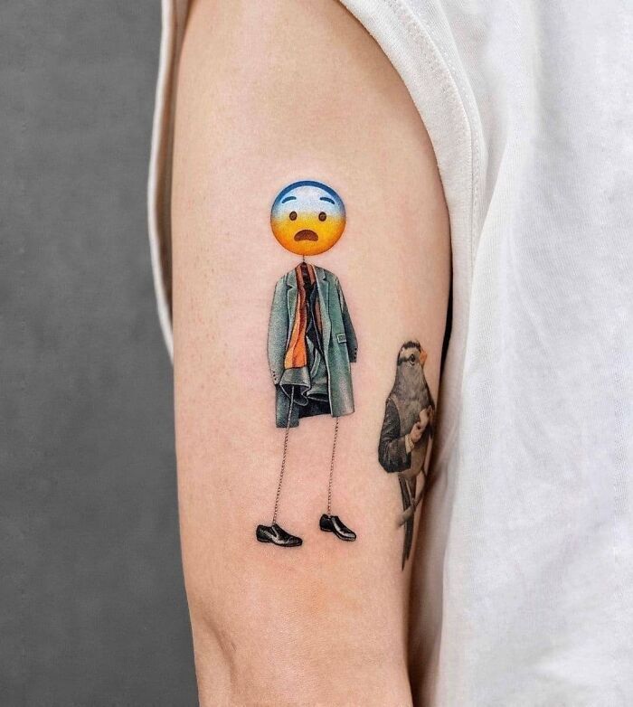 Tatuaje creativo en brazo con emoji cabeza desaprobando y cuerpo con abrigo y zapatos, ejemplo de tatuajes graciosos y originales.