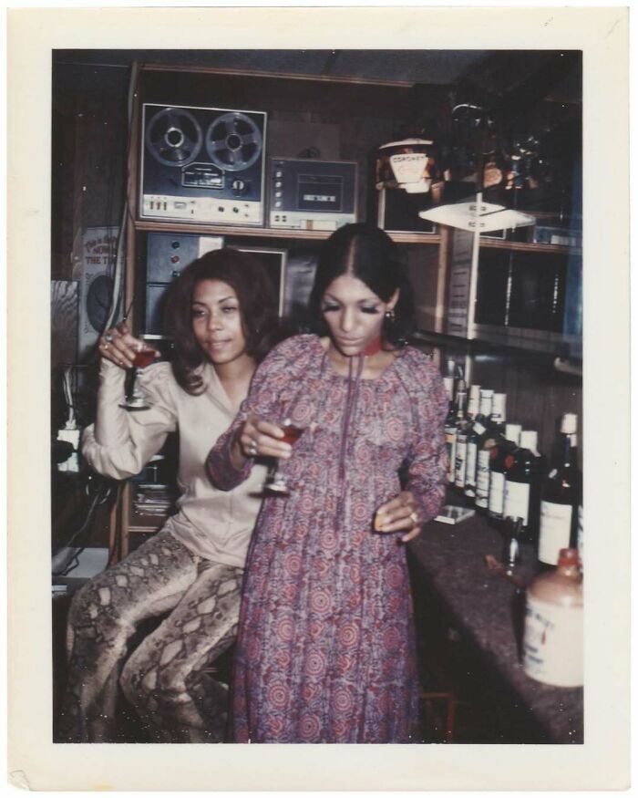 Two women in vintage attire enjoying drinks at a home bar with retro audio equipment in a mesmerizing vintage photo.