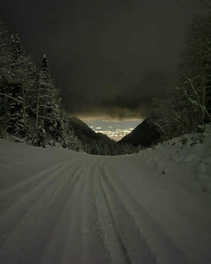 Snow-covered road disappearing between dark, empty spaces in a forested mountain setting, creating an unsettling yet fascinating scene.