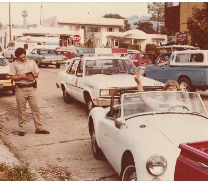 Vintage photo of a police officer writing a ticket while a woman in a convertible waves, capturing a moment in history.