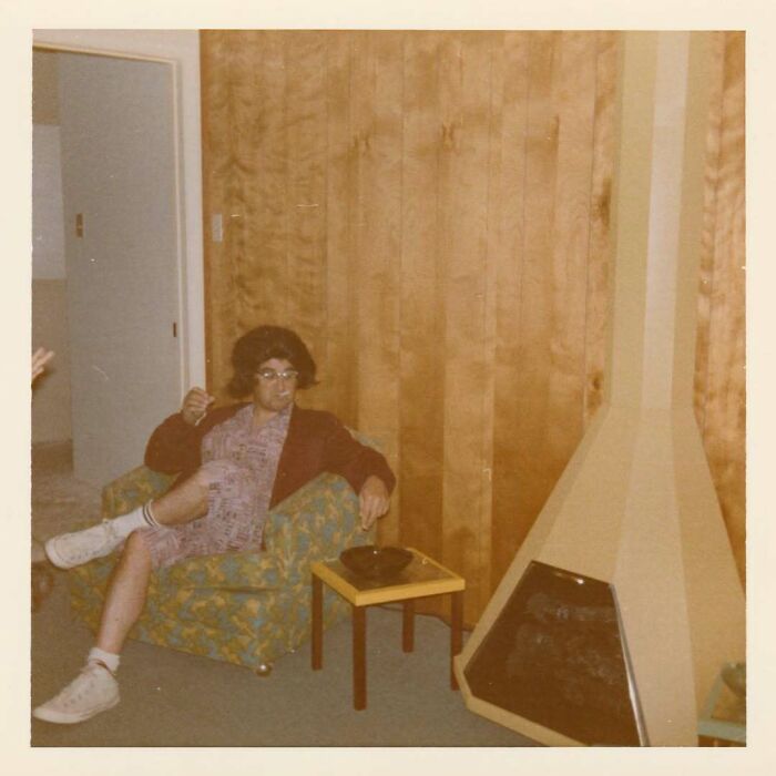 Vintage photo of a person sitting in a patterned armchair next to a retro fireplace in a wood-paneled room.