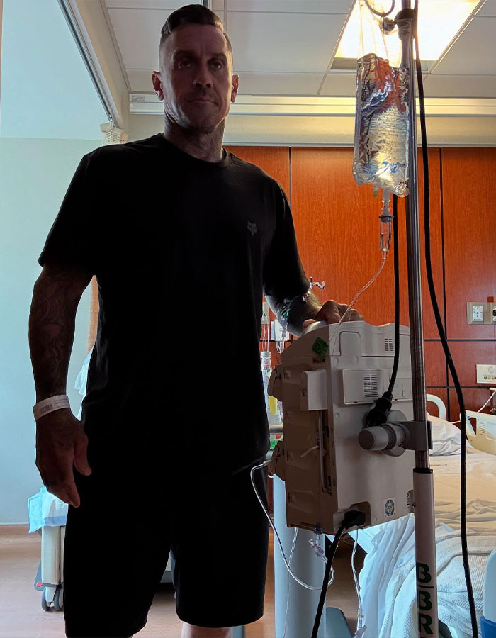 Pink's husband standing in hospital room next to IV drip after bike accident surgery, showing recovery progress.