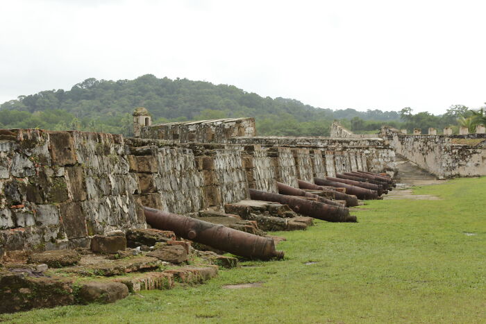 Ancient stone fortress wall with lined cannons amidst green grass and forested hills, a stunning UNESCO treasure site.