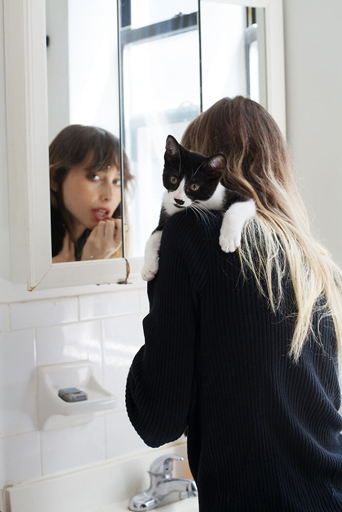 Black and white cat perched on a woman’s shoulder in front of a bathroom mirror, a candid cat photography moment.