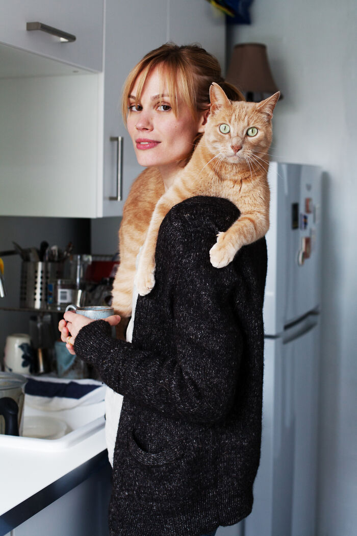 A woman in a black sweater holding a cup with an orange cat perched on her shoulder in cozy cat photography style.