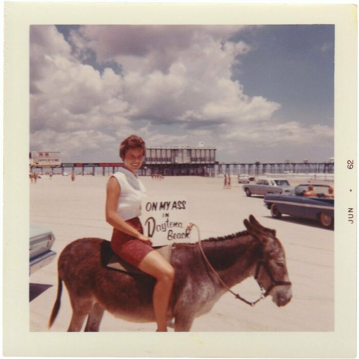 Vintage photo of a woman smiling on a donkey at Daytona Beach with cars and a pier in the background.