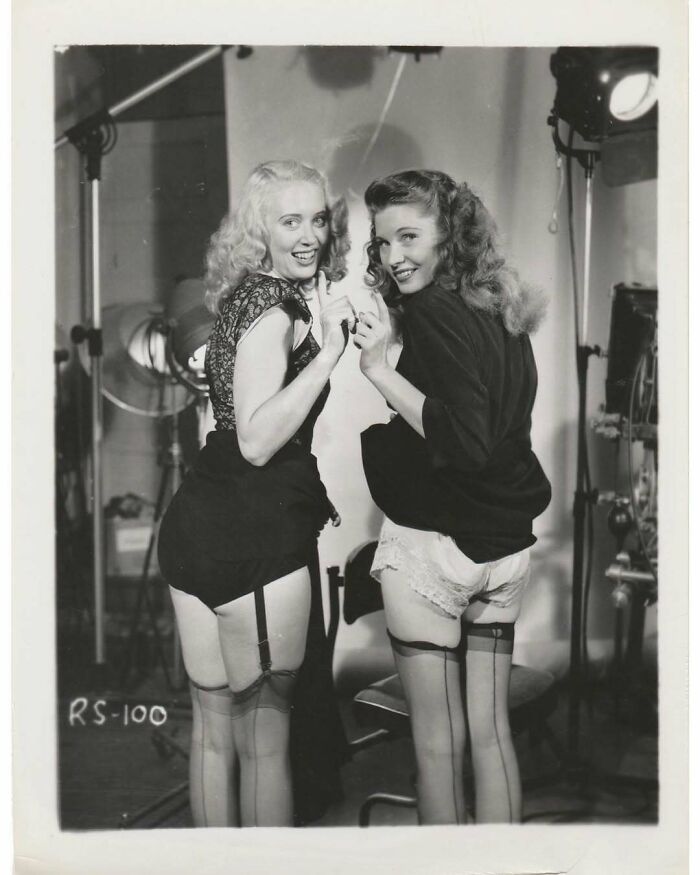 Two women in vintage stockings and garters posing playfully in a black and white vintage photo studio setting.