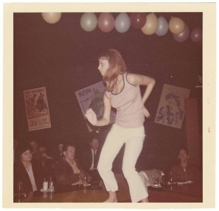 Young woman dancing on a table at a vintage party, surrounded by seated men and colorful balloons, classic vintage photo.