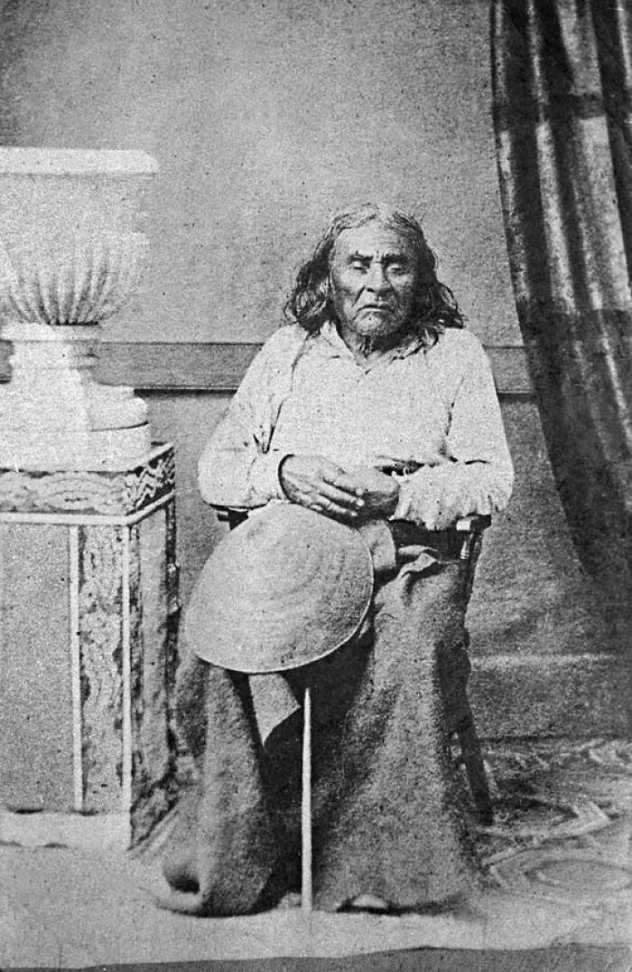 Vintage black and white photograph of an elderly Native American man seated holding a hat and walking stick, historical facts concept.
