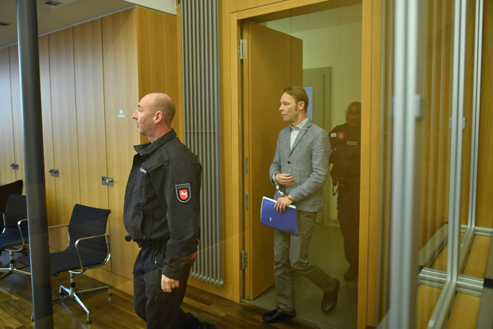 Man suspected in Madeleine McCann case smirks while escorted by security in a courtroom with wooden paneling.