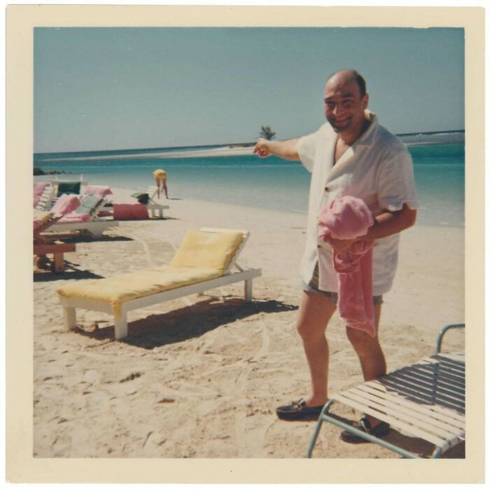 Man on a vintage beach pointing towards the ocean with lounge chairs and a person in the background on sandy shore.