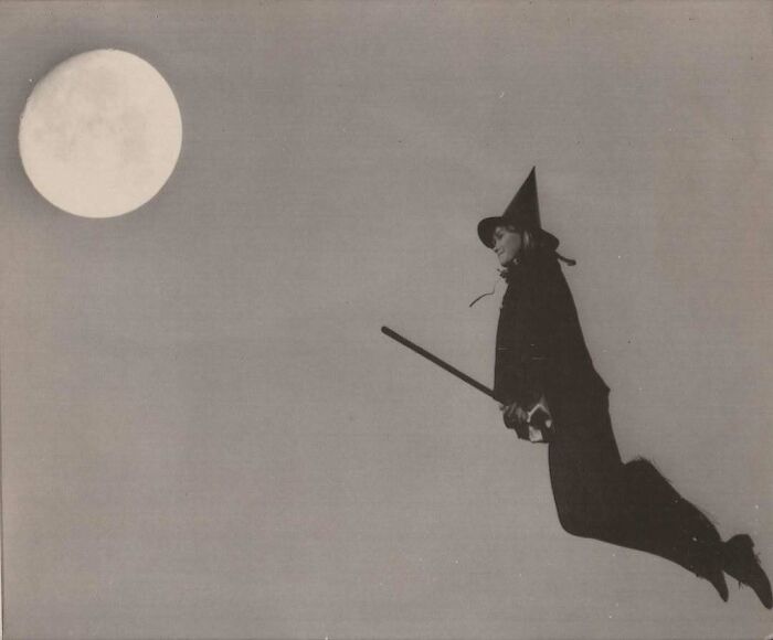 Vintage photo of a person dressed as a witch flying on a broomstick against a full moon night sky.