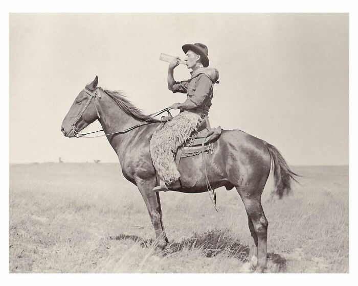 Vintage photo of a cowboy on horseback drinking from a bottle in an open field, showcasing captivating moments from history.