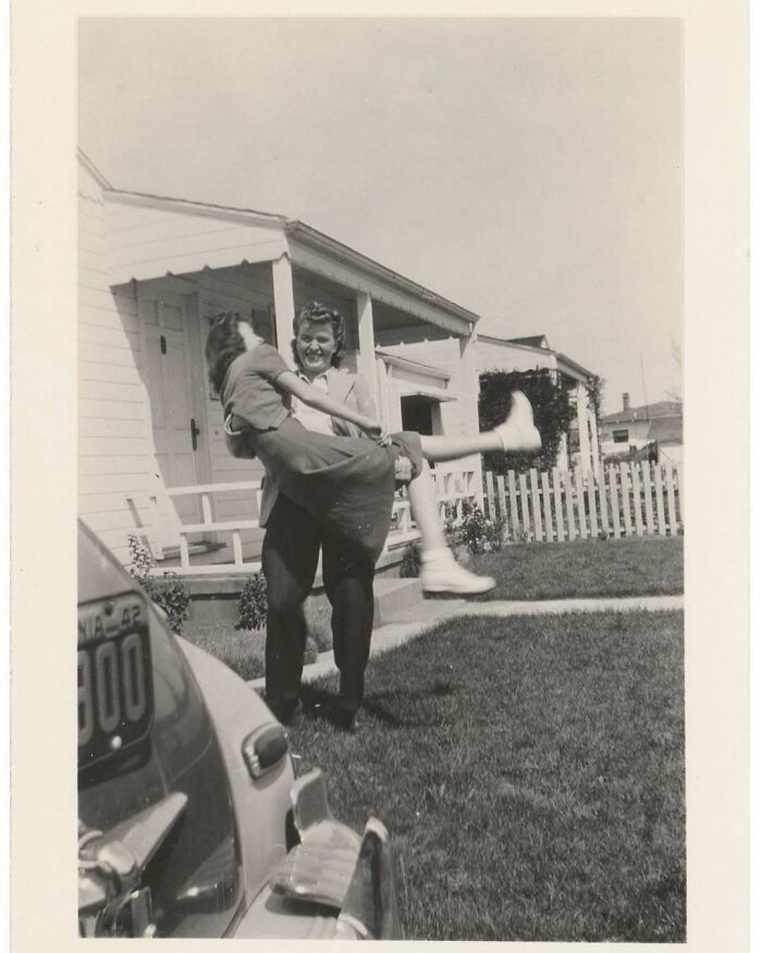 Vintage photo of a man joyfully lifting a woman outside a suburban home, capturing a charming moment in history.