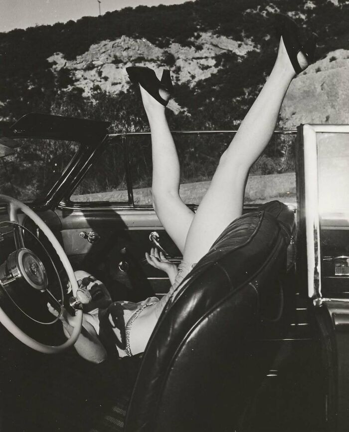 Vintage photo of a woman in high heels playfully posing with her legs raised inside a classic convertible car outdoors.