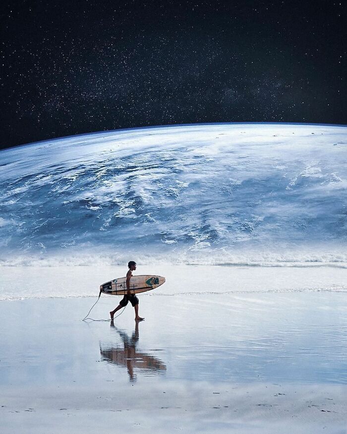 Surfer walking on a reflective beach with a large Earth photo manipulation and collage visible in the sky above.