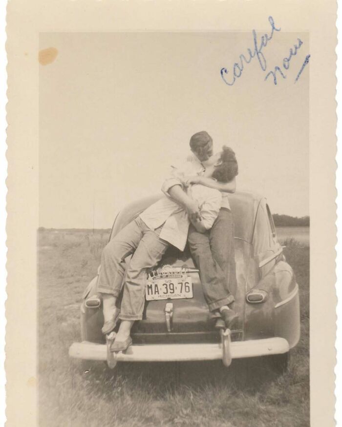 Vintage photo of a couple embracing on the back of a classic car in an open field, showcasing timeless romance and history.