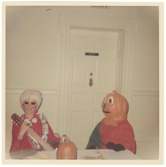 Vintage photo of two people in costumes, one playing guitar, beside a pumpkin on a table in front of a men’s restroom door.