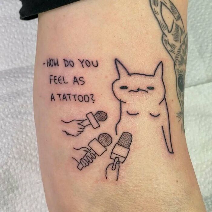 Tattoo of a cat being interviewed with multiple microphones and text asking how it feels as a tattoo, funny tattoo design.