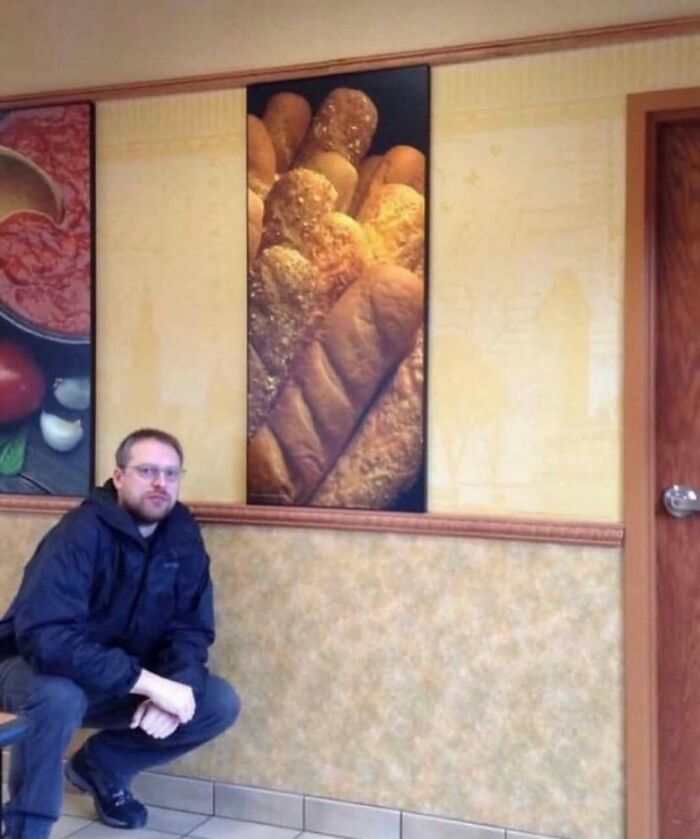 Man in a black jacket posing inside a restaurant with bread artwork on the wall, relatable memes context implied