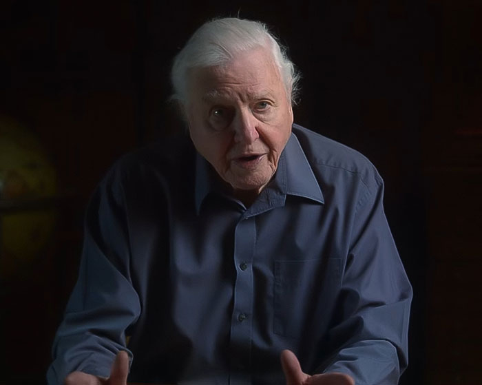 David Attenborough in a dark room wearing a blue shirt, speaking while sharing an urgent warning about life nearing its end.