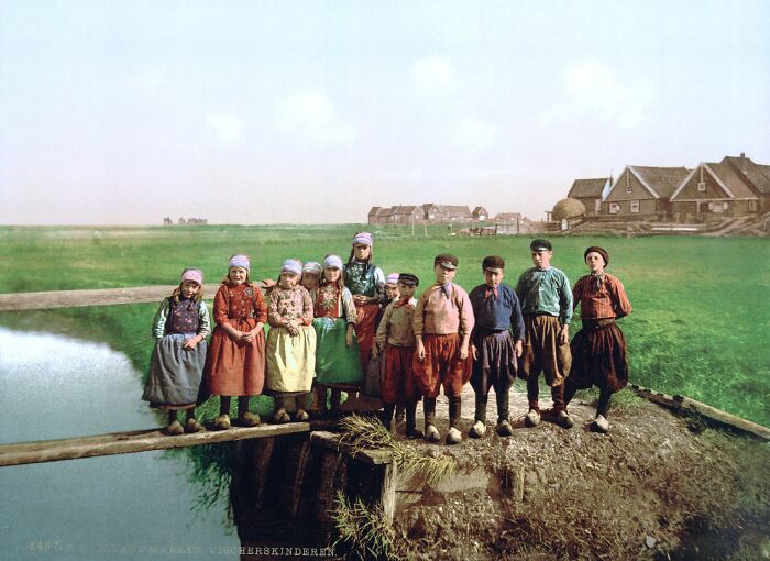 Group of children in traditional clothing standing outdoors near a pond in a colorized photo of cultural past