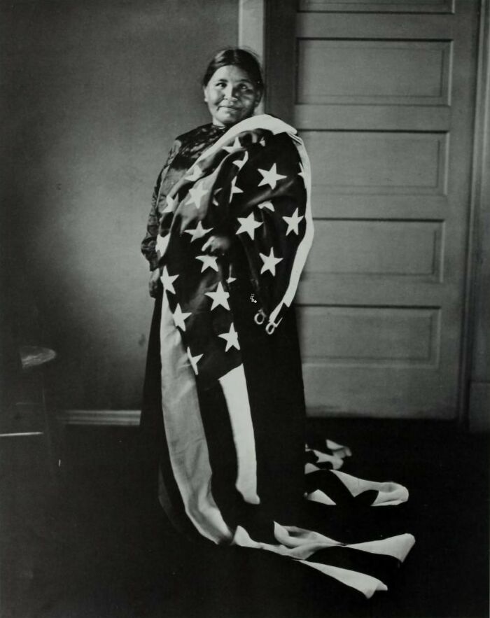 Native American woman wrapped in an American flag, symbolizing female strength in a time-weathered photograph.