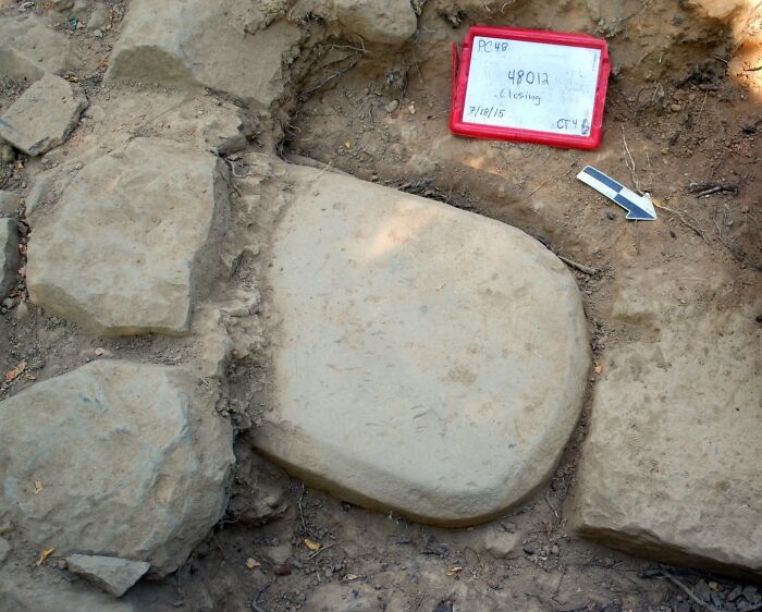 Ancient stone artifact partially unearthed at archeological site with markers indicating excavation details in soil.