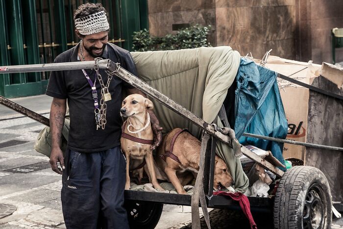 Homeless man standing beside a small cart with two dogs, capturing the lives of homeless people and their dogs.