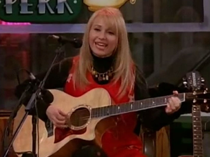 Friends star who sang Smelly Cat playing guitar and singing with a youthful glow on a TV show set.