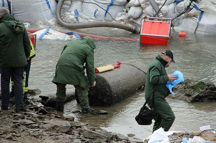 Experts in protective gear examining a large bizarre historical artifact partially submerged in water near the shore.
