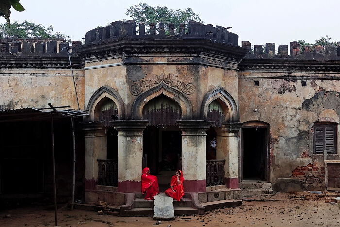 Old rustic building in Simultala with two women in red sarees sitting and chatting in a timeless escape setting.