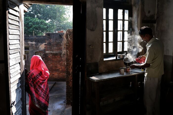 Man pouring steaming tea inside a rustic kitchen while a woman in a red saree stands near an open door in Simultala.