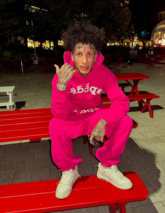 Island boy sitting on red bench at night, wearing bright pink outfit and showing tattoos on face and hands.