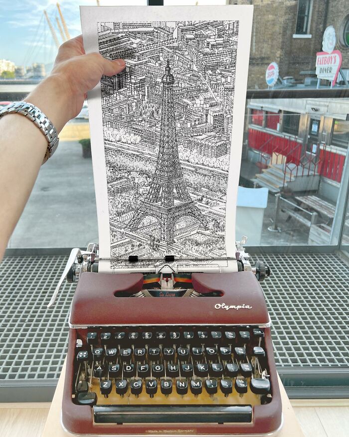 Hand holding detailed typewriter drawing of Eiffel Tower above a vintage Olympia typewriter on a table near window.