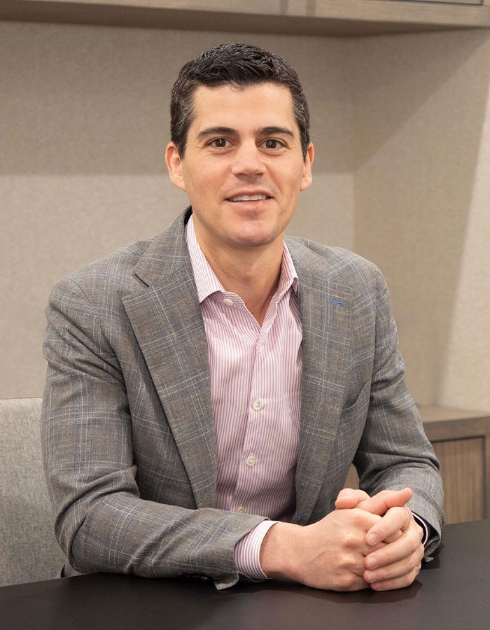 Plastic surgeon in a gray blazer and pink striped shirt, smiling and seated at a desk in a professional office setting Plastic surgeon in a gray blazer and pink striped shirt, smiling and seated at a desk in a professional office setting