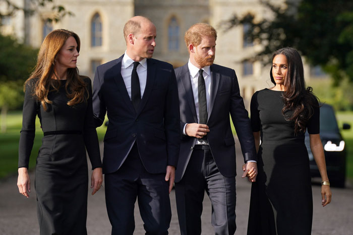 Meghan Markle and Kate Middleton in black outfits walking with Prince William and Prince Harry at a formal outdoor event.