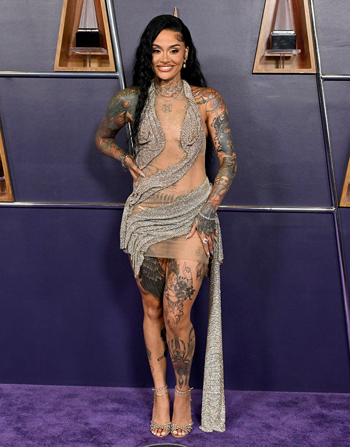 Woman with extensive tattoos wearing a sheer glittery dress, posing on the purple carpet at the worst dressed stars event.