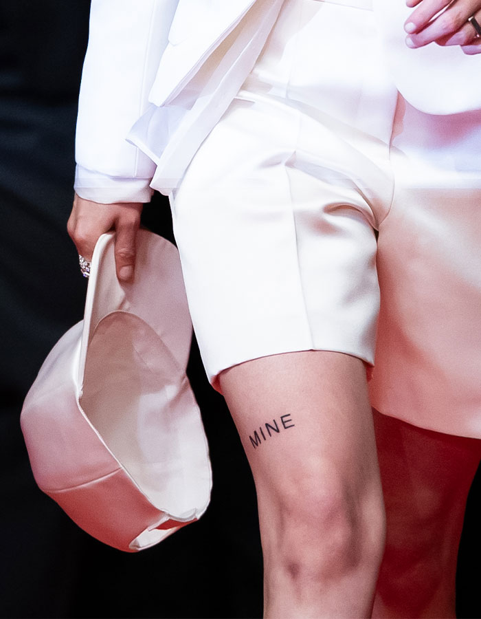 Close-up of Kristen Stewart's thigh tattoo with word Mine, wearing white shorts and holding a matching white hat.