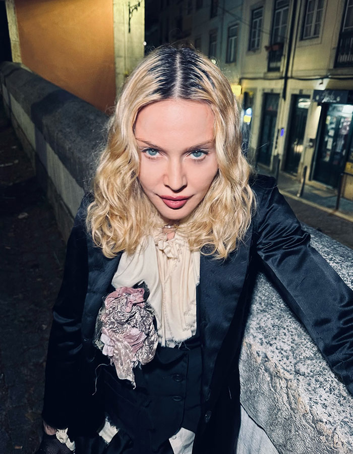 Blonde woman standing on a stone bridge at night, wearing a black jacket and blouse with a large floral brooch.