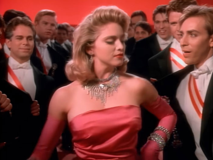 Madonna in a glamorous red dress and jewelry at a formal event, surrounded by men in tuxedos with sashes.