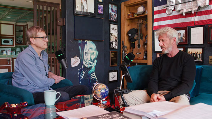 Two men in a podcast studio, one speaking into a microphone, the other listening, with Sean Penn among them.