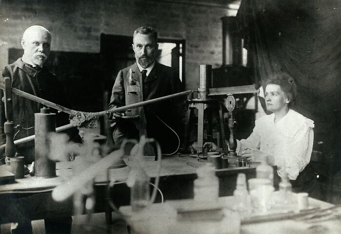 Marie Curie in a historic laboratory setting with two men, surrounded by early scientific equipment and apparatus.