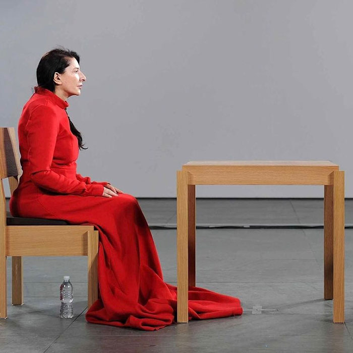Controversial artist in red dress sitting on wooden chair next to table in public gallery detailing her experience. Controversial artist in red dress sitting on wooden chair next to table in public gallery detailing her experience.