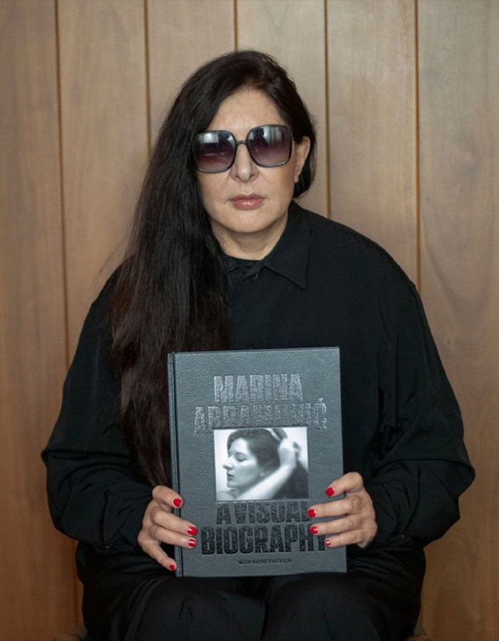 Controversial artist wearing sunglasses, holding a visual biography book, seated against a wooden panel background. Controversial artist wearing sunglasses, holding a visual biography book, seated against a wooden panel background.