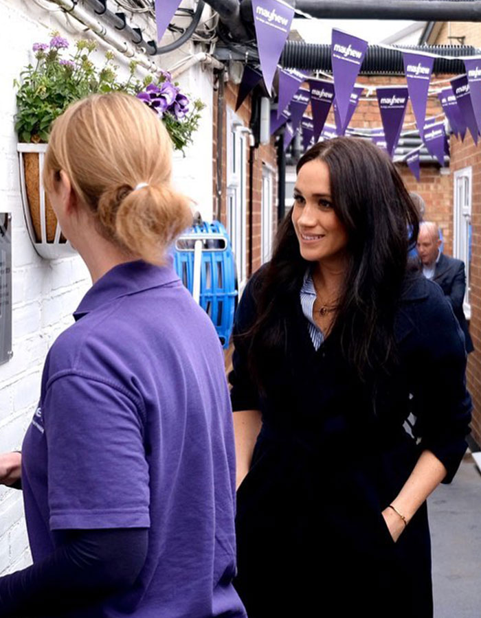 Meghan Markle smiling and talking with a woman in a purple shirt under purple Mayhew banners in a narrow alley.